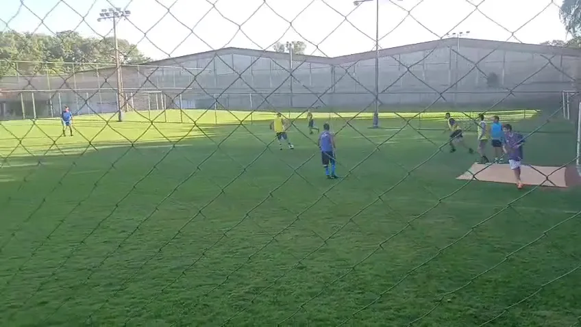 Jogo de futebol society em grama natural visto pelo alambrado, no Campo do Morjube